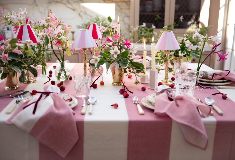 Decorative table setting on pink stripe linen tablecloth, napkins, and floral arrangements and brass tealight candle stands with striped shades