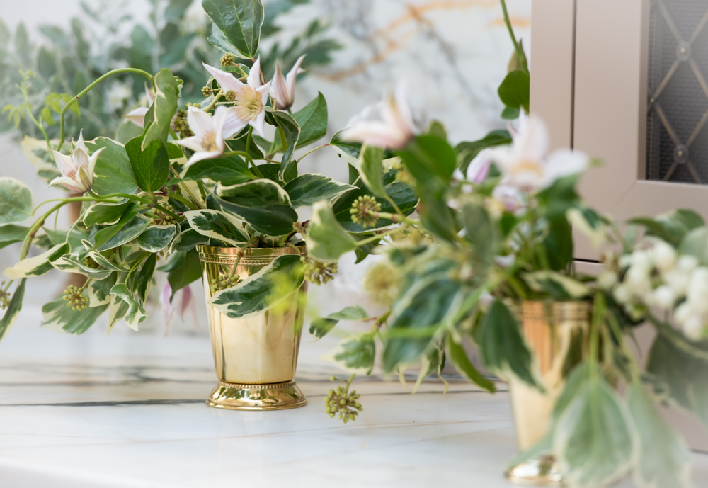 Two brass julep cup vases with green and white flower arrangements on a marble counter