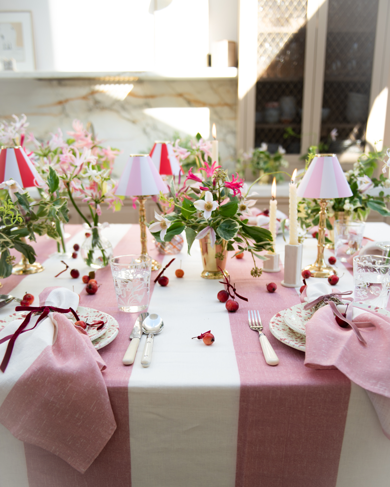 Elegant table setting with pink and white stripe linen tablecloth and napkins, floral centerpieces in brass vases and red, white and pink stripe candle shades on brass tealight stands