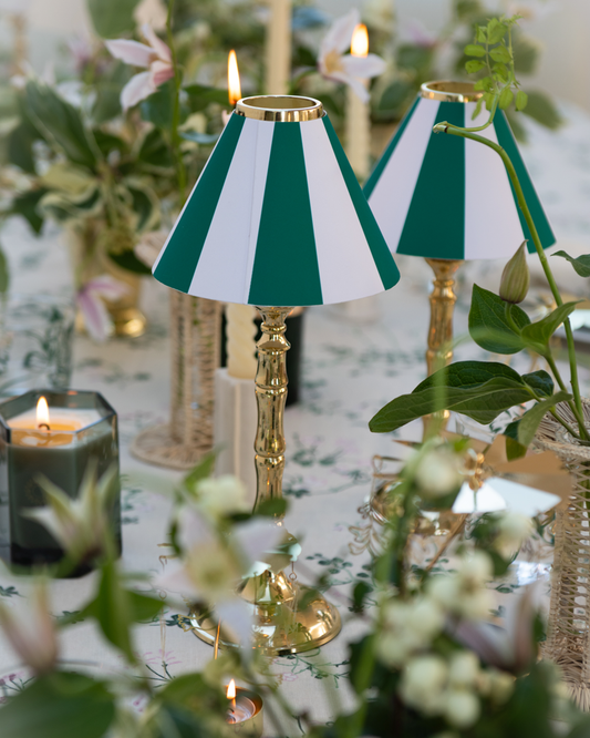 Two green and white striped candle shades on brass tealight candle stands surrounded by floral arrangements and scented candles on botanica linen tablecloth
