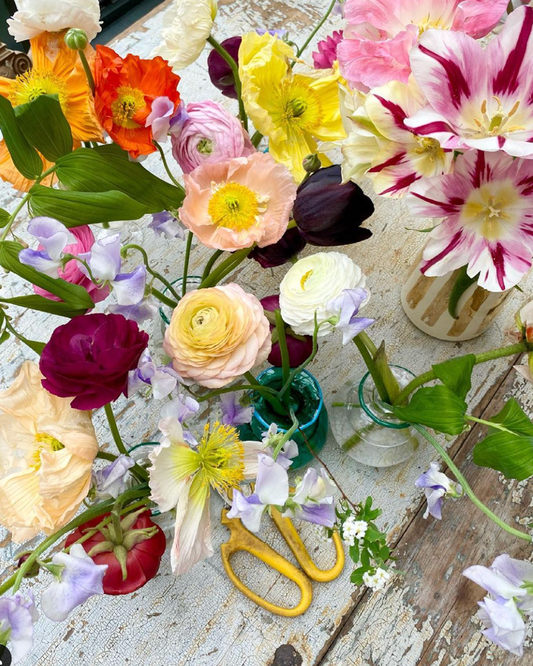 Colorful bud vases of flowers on a rustic wooden table with a pair of yellow scissors.